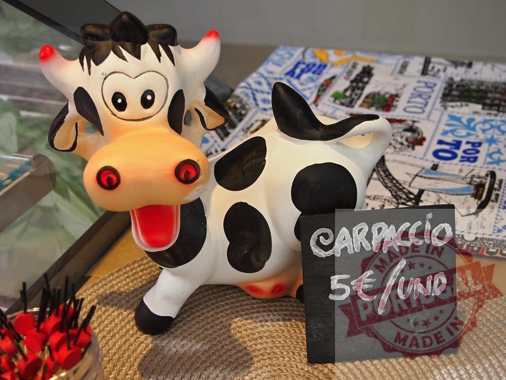 Ceramic cow figurine advertising carpaccio price Porto | Ceramic cow figurine standing next to a chalkboard sign with carpaccio price in a shop - Realisiert mit Pictrs.com
