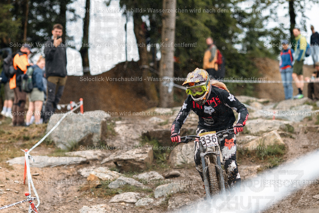Mountainbiker im Sturmlauf | Ein Mountainbiker rast einen staubigen, felsigen Pfad hinunter, während er einen Sprung vorbereitet. Der Fahrer trägt einen schwarzen Helm, eine Schutzkleidung und die Nummer 336 auf seinem Rücken. Im Hintergrund sind Zuschauer und Bäume zu sehen. - Realisiert mit Pictrs.com