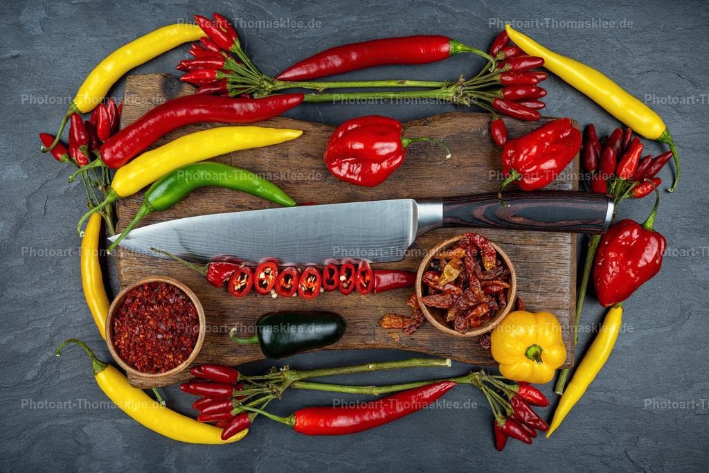 Bunte Chilis und Paprika auf Holzbrett mit Messer | Auf einem rustikalen Holzbrett liegen verschiedene Chilis und Paprika in lebendigen Farben. Rote, gelbe und grüne Schoten umrahmen das Brett kreisförmig auf dunklem Schieferuntergrund. Ein scharfes Küchenmesser liegt mittig auf dem Holz, neben kleinen Schalen mit getrockneten Chilis und Chilipulver. Die Komposition zeigt die Vielfalt scharfer Gewürze in ihrer natürlichen Schönheit. Frische und getrocknete Varianten ergänzen sich harmonisch in diesem appetitlichen Arrangement für die anspruchsvolle Küche. - Realisiert mit Pictrs.com