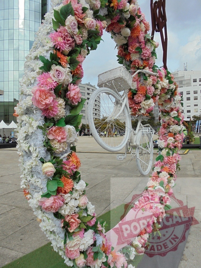 Floral heart arch framing white bicycle art installation | Large heart shape arch with colorful flowers framing a white bicycle in an outdoor plaza - Realisiert mit Pictrs.com