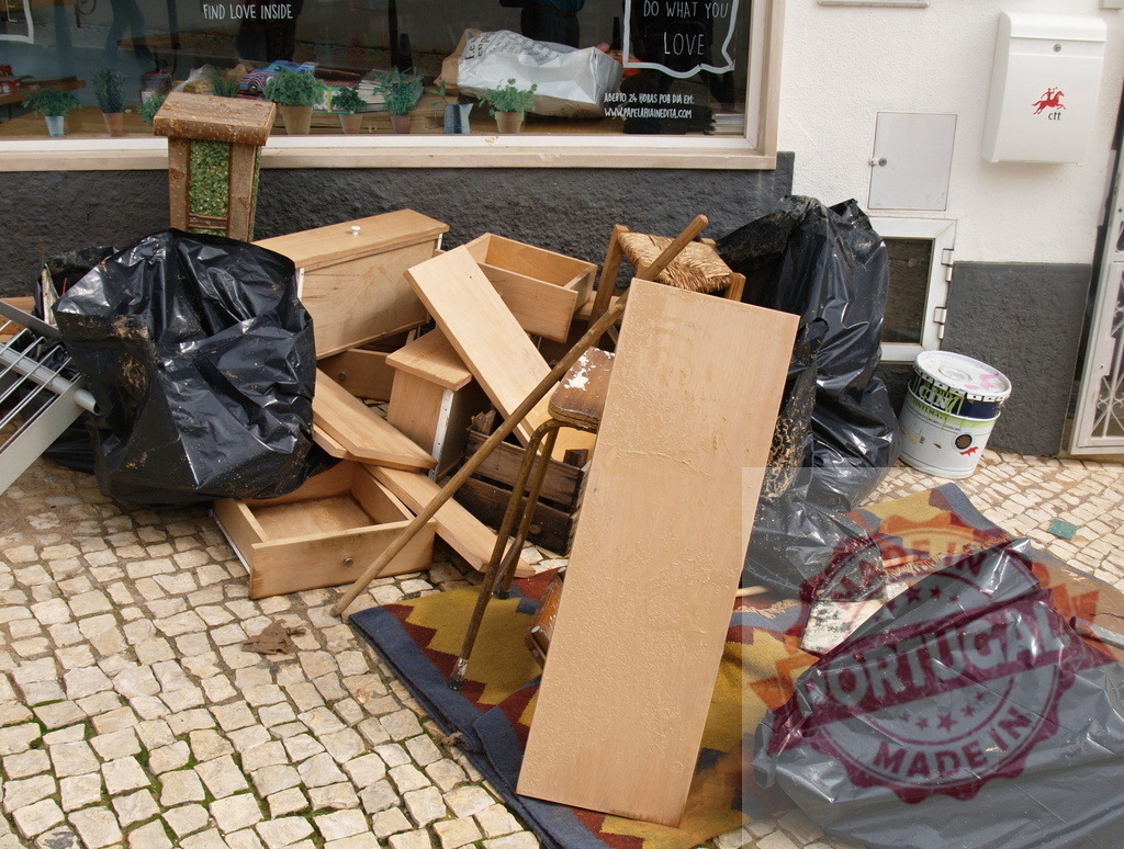 Discarded furniture and rubbish piling on a cobbled street | Albufeira Flood - discruction by weather problems Waste materials and black bags filling a street after a renovation or cleanup - Realized with Pictrs.com