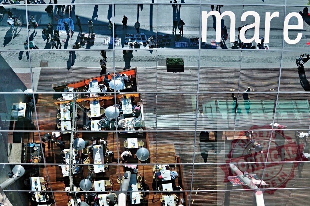Urban reflection showing people dining and walking on street. Spain - Barcelona | Glass facade reflecting people walking on the street above an outdoor restaurant - Realized with Pictrs.com