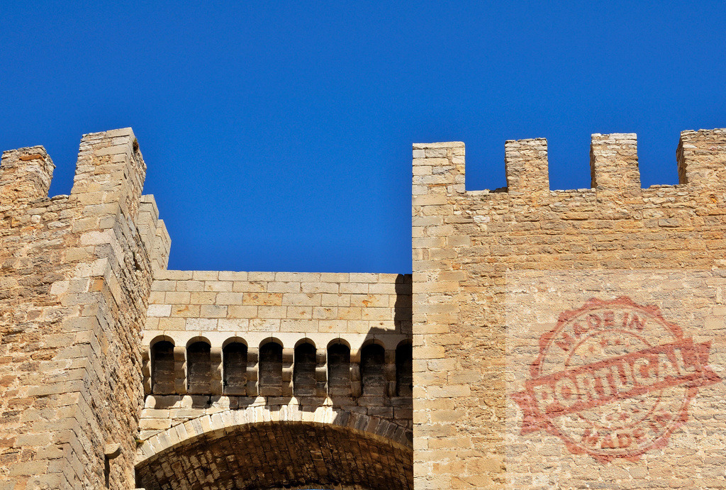 Medieval castle wall crenellations against blue sky. Spain - Castellon | Stone fortress wall with battlements, arch, and machicolations under a clear blue sky - Realized with Pictrs.com