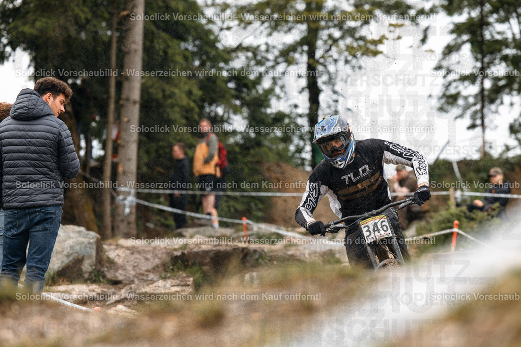 Mountainbike-Action im Wald | Das Foto zeigt einen Mountainbiker in voller Montur, der einen staubigen, kurvigen Pfad in einem bewaldeten Gebiet durchquert. Der Biker trägt einen blauen Helm und eine schwarze Jacke mit dem Schriftzug 'TLD'. Er fährt mit hoher Geschwindigkeit und ist in der Mitte des Bildes zu sehen. Links vom Biker steht ein Zuschauer in einer blauen Jacke und Jeans, der das Rennen beobachtet. Im Hintergrund sind weitere Zuschauer und Bäume zu sehen. Der Pfad ist mit Staub bedeckt und weist einige Hindernisse auf. - Realisiert mit Pictrs.com