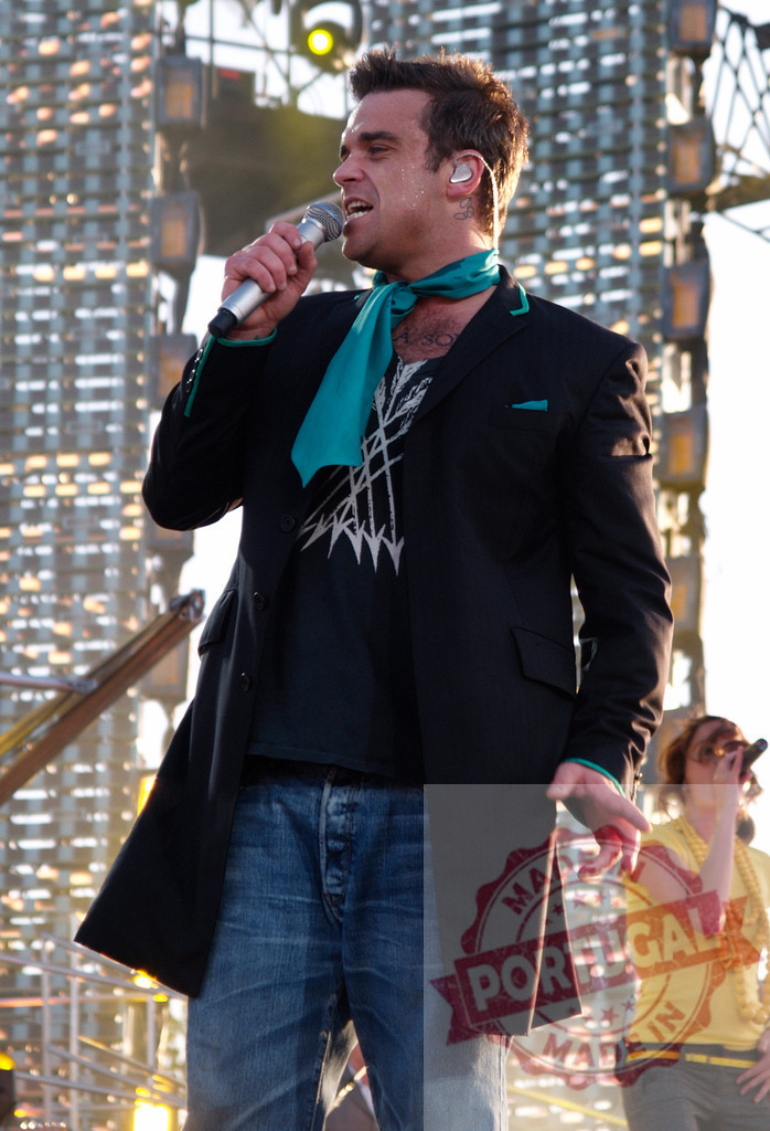 Robbie Williams singing during a concert, wearing a black jacket and turquoise scarf | Robbie Williams performing on stage, holding a microphone and wearing a turquoise scarf - Realisiert mit Pictrs.com