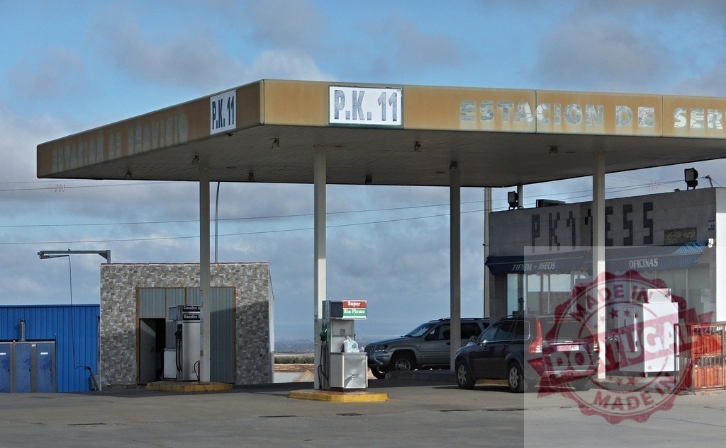 Roadside gas station with cars parked and blue sky | Gas station with old sign and parked cars under a weathered canopy on a cloudy day - Realisiert mit Pictrs.com