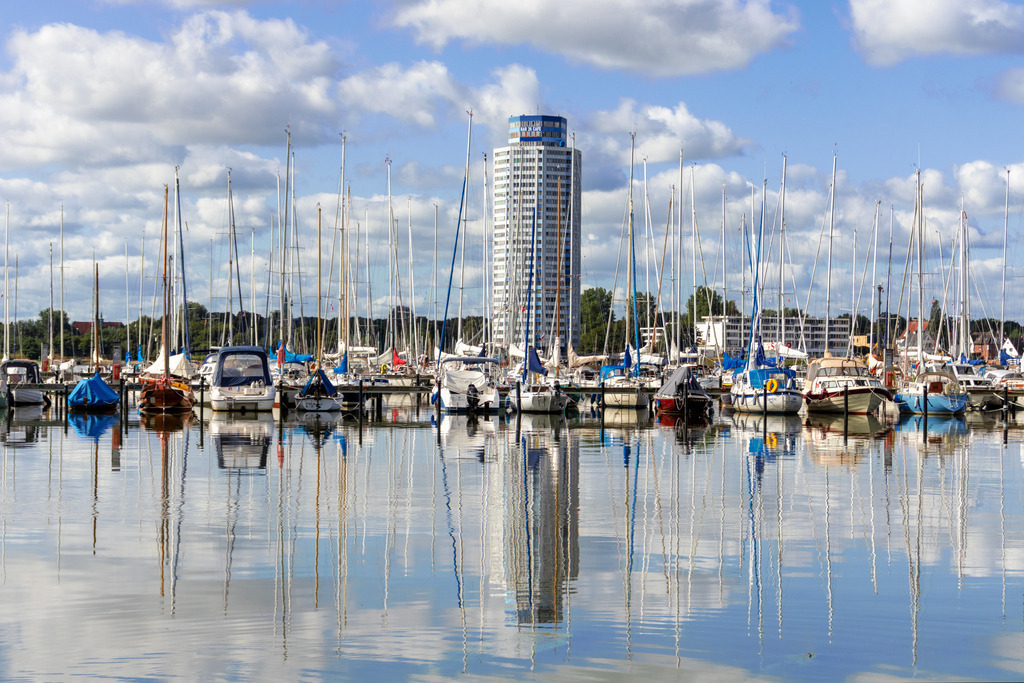 Wandbild: Yachthafen in Schleswig mit schöner Spiegelung | Dieses Wandbild im Querformat zeigt den Yachthafen in Schleswig. Die Segelboote sowie der Wikingturm und die Wolken spiegeln sich auf dem Wasser der Schlei - Realisiert mit Pictrs.com