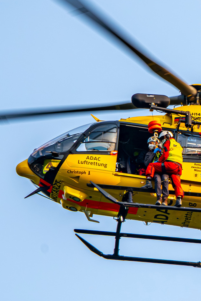 Bergretter | Ein markanter gelber ADAC-Rettungshubschrauber (Christoph 1) befindet sich im Schwebeflug vor einem klaren, hellen Himmel. An der Seite ist die Rettungswinde im Einsatz: Ein Luftretter sichert eine Person, während sie gemeinsam am Seil unterhalb der offenen Kabinentür hängen – ein intensiver Einblick in die lebensrettende Arbeit der Bergwacht oder Luftrettung. - Realisiert mit Pictrs.com