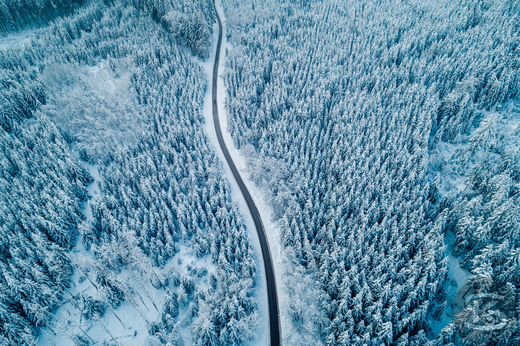 Winterwald aus der Luft – Verschneite Straße durch gefrorenen Nadelwald | Diese Drohnenaufnahme zeigt eine kurvenreiche Straße, die sich durch einen dicht verschneiten Nadelwald schlängelt. Aus der Vogelperspektive wird die geometrische Struktur sichtbar: Die organische Linie der Straße bildet einen starken Kontrast zu den vertikalen Mustern der schneebedeckten Bäume. Die kalten Blautöne verstärken die winterliche Atmosphäre und verleihen dem Wald eine fast abstrakte Qualität. - Realisiert mit Pictrs.com