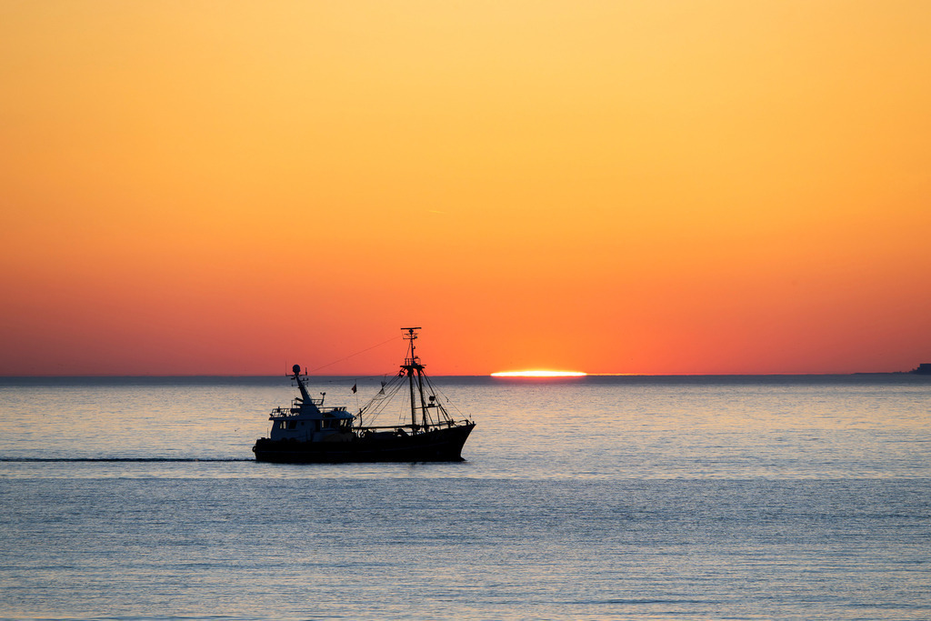 Sonnenuntergang am Meer | Das Foto fängt einen stimmungsvollen Moment auf dem Meer ein. Ein Fischkutter ist als dunkle Silhouette vor einem intensiven Sonnenuntergang zu sehen. Die Sonne ist fast vollständig hinter dem Horizont verschwunden und hinterlässt einen leuchtend orange-gelben Farbverlauf am Himmel, der sich sanft im ruhigen Wasser spiegelt. - Realisiert mit Pictrs.com