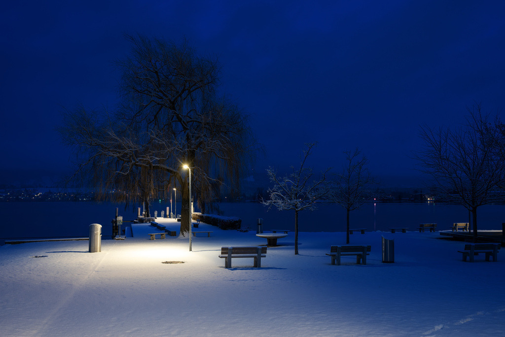 Ländte Hünibach | Eine Langzeitbelichtung fängt die ruhige Winterlandschaft der Uferpromenade von Huenibach ein. Eine markante Strassenlaterne beleuchtet den frisch gefallenen Schnee und kontrastiert mit den tiefblauen Farbtönen des Nachthimmels und den ruhigen Gewässern des Thunersees. Kahle Bäume, schneebedeckte Bänke und entfernte Lichter am gegenüberliegenden Ufer vervollständigen die friedliche Szene. - Realisiert mit Pictrs.com