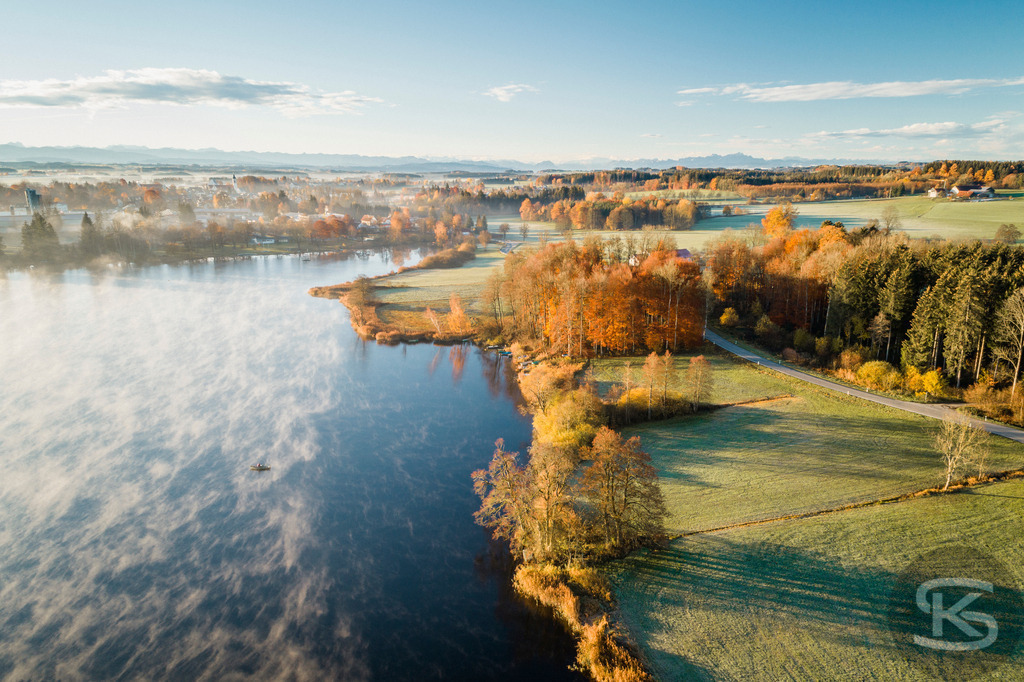 Herbstlandschaft mit Nebel über See - Luftaufnahme Bayern im Morgenlicht | Spektakuläre Drohnenaufnahme einer herbstlichen Seelandschaft im warmen Morgenlicht. Mystischer Bodennebel über dem Wasser, goldene Herbstbäume und verschneite Alpen im Hintergrund vereinen sich zu einem malerischen Panorama in Süddeutschland. - Realisiert mit Pictrs.com