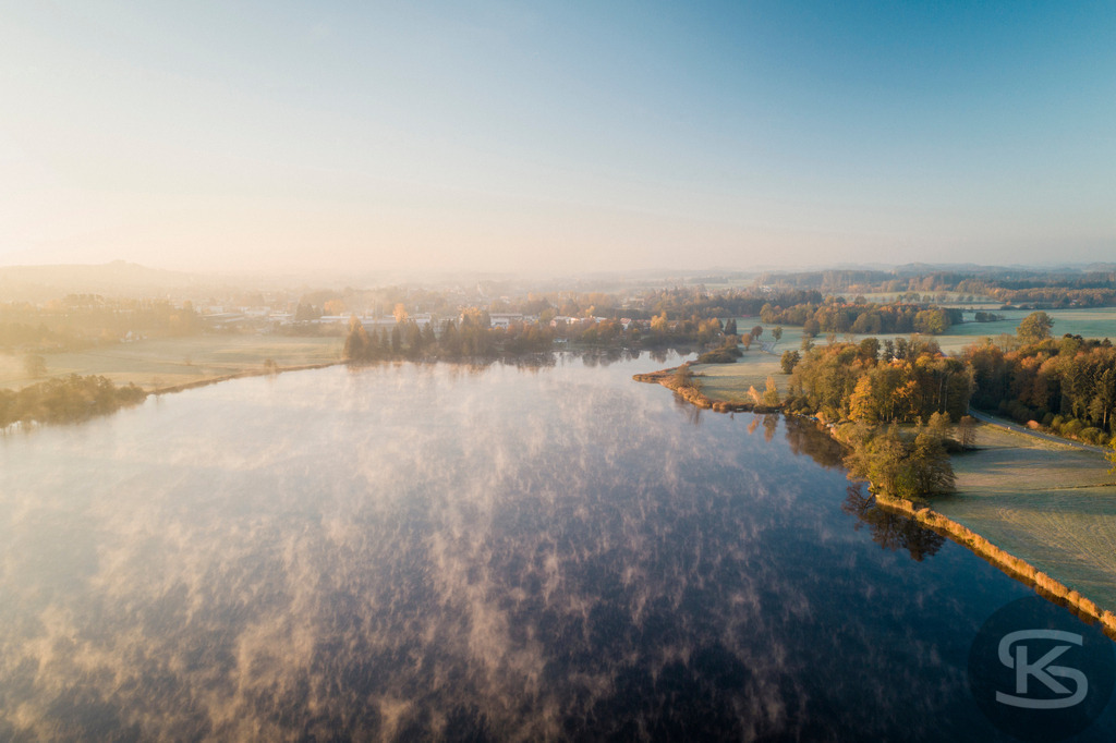 Herbstlandschaft mit Nebel über See - Luftaufnahme Bayern im Morgenlicht | Spektakuläre Drohnenaufnahme einer herbstlichen Seelandschaft im warmen Morgenlicht. Mystischer Bodennebel über dem Wasser, goldene Herbstbäume und verschneite Alpen im Hintergrund vereinen sich zu einem malerischen Panorama in Süddeutschland. - Realisiert mit Pictrs.com