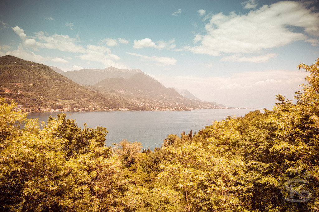 Idyllischer Panoramablick auf den Gardasee | Ein malerischer Blick auf den Gardasee in Italien, umgeben von majestätischen Bergen und üppiger, warmer Vegetation. Das Bild fängt die friedliche Atmosphäre der Region mit ihren kleinen Küstensiedlungen am Ufer ein und lädt zum Träumen von Urlaub und mediterranem Flair ein. - Realisiert mit Pictrs.com