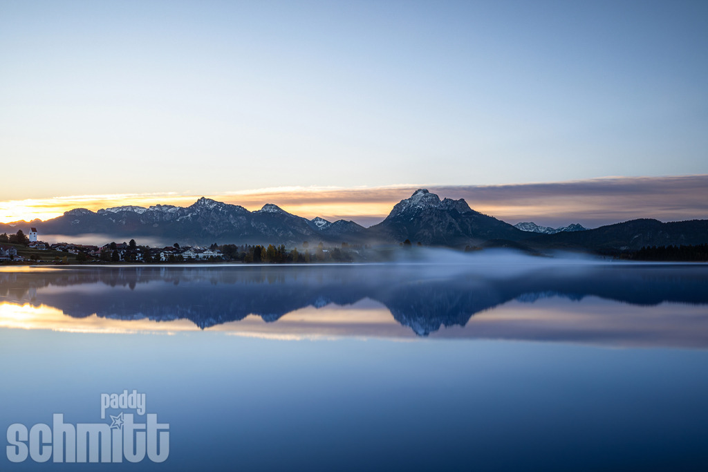 Forggensee bei Sonnenaufgang mit Wasserspiegelung und Bergpanorama | Stimmungsvoller Sonnenaufgang am Forggensee, bei dem sich die ersten warmen Lichtfarben und das imposante Bergpanorama im ruhigen Wasser spiegeln. Eine idyllische Aufnahme mit der besonderen Atmosphäre eines frühen Morgens im Ostallgäu. - Realisiert mit Pictrs.com