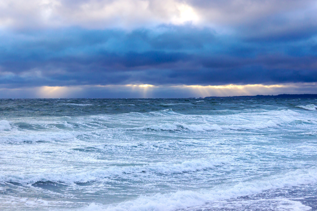 Wandbild: Sonnenschein, Wellen und Wolken | Dieses Wandbild im Querformat zeigt eine schöne Lichtstimmung am Strand in Booknis. Auf der Ostsee sind zahlreiche Wellen mit Schaumkronen zu sehen. Am Himmel befinden sich dunkle Wolken, die durch die morgendliche Lichtstimmung in vielen verschiedenen Farben leuchten. Durch die dunklen Wolken gelangen Sonnenstrahlen bis zum Meer.  - Realisiert mit Pictrs.com