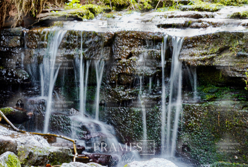 Kleiner Wasserfall | Das Foto zeigt einen kleinen Wasserfall, der über recht gerade, übereinanderliegende Steinstrukturen herabfließt und sich in viele schmale Wasserläufe aufteilt. Durch die lange Verschlusszeit erscheint das herabfließende Wasser als weicher, weißer, fast seidiger Strahl, der einen starken Kontrast zu den dunkleren Felsen bildet. Die Aufnahme betont die gleichmäßige Form des Wasserfalls, die klaren Linien der Steine und die ruhige, atmosphärische Wirkung der Langzeitbelichtung. Das Zusammenspiel aus Struktur, Bewegung und weichem Licht verleiht der Szene eine beruhigende und zugleich dynamische Ästhetik. - Realisiert mit Pictrs.com