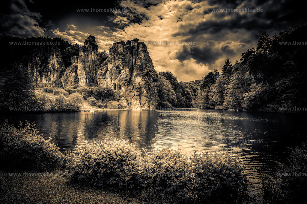 Externsteine – Zeitlose Felsen in dunklem Sepia | Dies ist eine wahrhaft beeindruckende Kunstfotografie der Externsteine, eines der faszinierendsten Naturdenkmäler Deutschlands, das tief im Teutoburger Wald im Bundesland Nordrhein-Westfalen liegt. Das Bild wurde in einem kräftigen, dramatischen Sepia-Ton bearbeitet, der alle ablenkenden Farbtöne entfernt und durch etwas weitaus Emotionaleres ersetzt – eine zeitlose, fast eindringliche Atmosphäre, die eher an ein Gemälde als an eine Fotografie erinnert.Die Felsformation selbst erhebt sich aus dem Mittelgrund wie etwas Uraltes und Unbeugsames. Diese Sandsteinsäulen stehen seit Millionen von Jahren und tragen ein enormes historisches und kulturelles Gewicht. In vorchristlicher Zeit als Kultstätte genutzt und später von den frühen Christen, die Kapellen und Reliefs direkt in den Stein meißelten, waren die Externsteine schon immer ein Ort, der die Menschen innehalten und nachdenken lässt. Dieses Gefühl tiefer Geschichte kommt in diesem Bild deutlich zum Ausdruck – die Sepia-Bearbeitung verleiht der Szene etwas, das sie wirklich alt wirken lässt, als blicke man auf einen Moment aus einem anderen Jahrhundert.Der See im Vordergrund erstreckt sich weit und ruhig, seine Oberfläche fängt die warmen Goldtöne des Himmels ein und spiegelt die Felsen in einem sanften, schimmernden Spiegel wider. Niedrige Sträucher und Wildpflanzen säumen das nahe Ufer und verleihen dem Bild Struktur und ein Gefühl von natürlichem Wildwuchs. Hinter und um die Felsen herum füllt ein dichter Wald das Bild aus, dessen dunkle Umrisse von beiden Seiten herandrängen. - Realisiert mit Pictrs.com