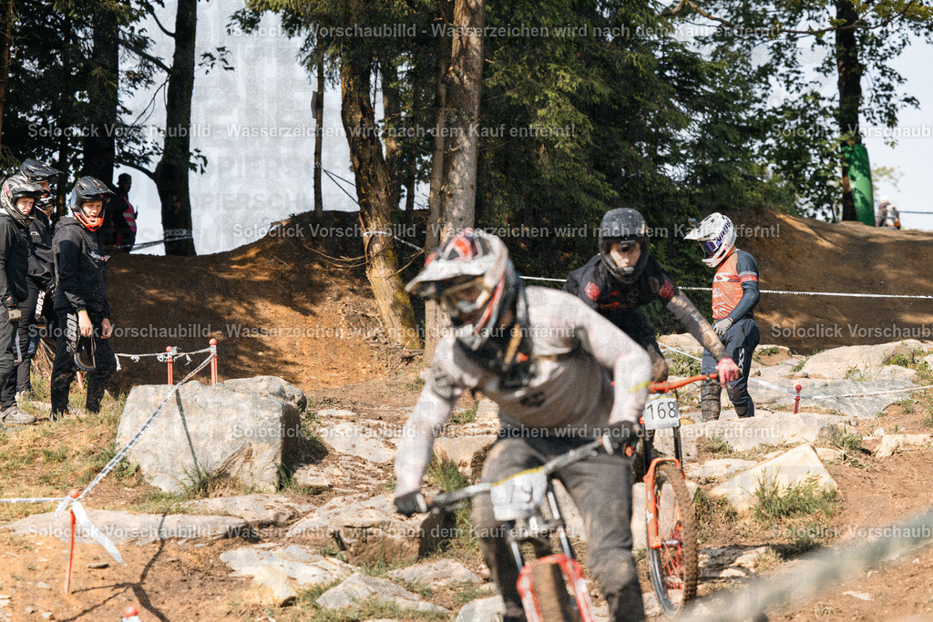 Mountainbike-Action im Gelände | Das Foto zeigt einen Mountainbiker in voller Ausrüstung, der mit hoher Geschwindigkeit einen anspruchsvollen, steinigen Pfad durch ein bewaldetes Gelände bewältigt. Im Hintergrund sind weitere Mountainbiker zu sehen, die ebenfalls den Kurs befahren. Die Szene vermittelt ein Gefühl von Geschwindigkeit, Herausforderung und Abenteuer. - Realisiert mit Pictrs.com
