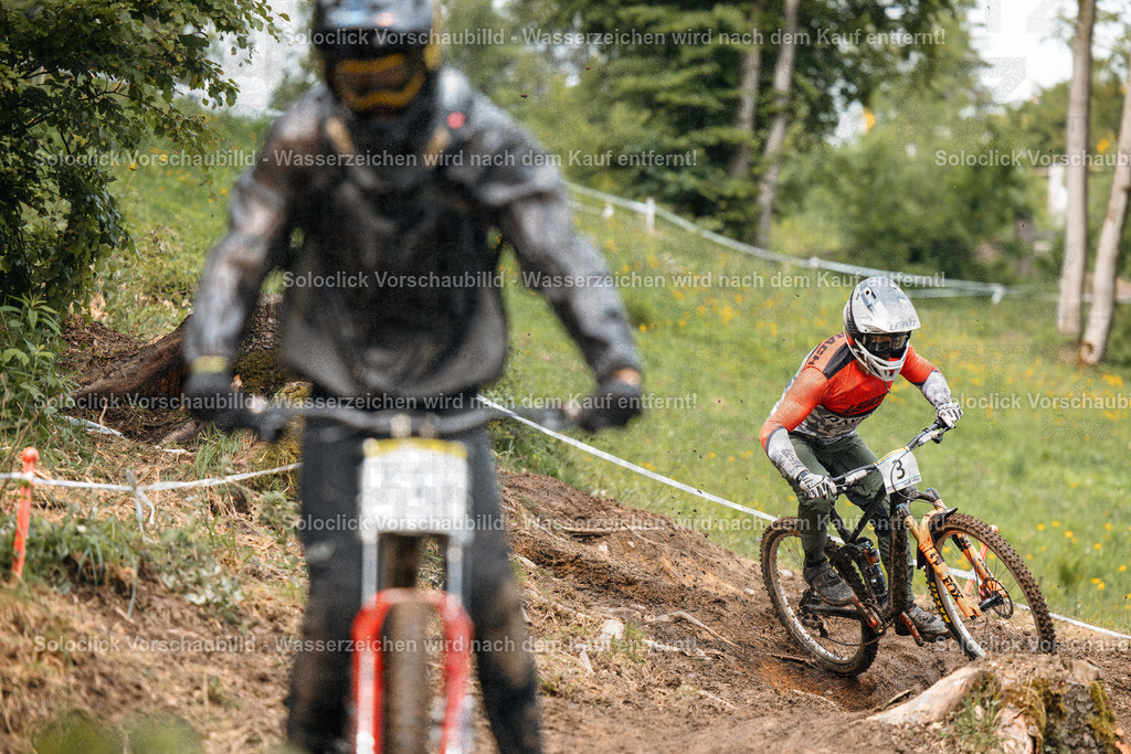 Mountainbike-Action | Das Foto zeigt zwei Mountainbiker in voller Montur, die einen anspruchsvollen Trail hinunterfahren. Der Vordergrundbiker ist in voller Fahrt und trägt einen roten Helm und eine orangefarbene Jacke. Er trägt die Nummer 3 auf seinem Rücken. Der Hintergrundbiker ist ebenfalls in Bewegung und trägt einen schwarzen Helm und eine Jacke. Der Trail ist staubig und kurvig, mit Gras und Büschen am Rand. Das Wetter ist bewölkt. - Realisiert mit Pictrs.com