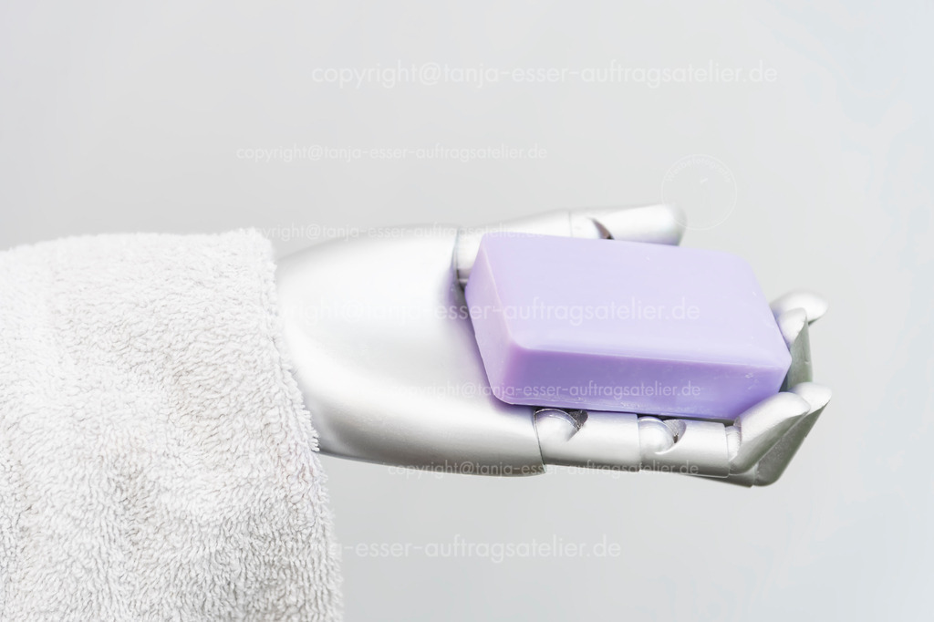 Robot takes care of people | Der Roboterarm hält Frotteehandtücher und ein Stück Seife in der Hand. Das Bild symbolisiert die Pflege von Menschen durch Maschinen. Konzeptuelles Foto vor weißem Hintergrund mit Copy Space.