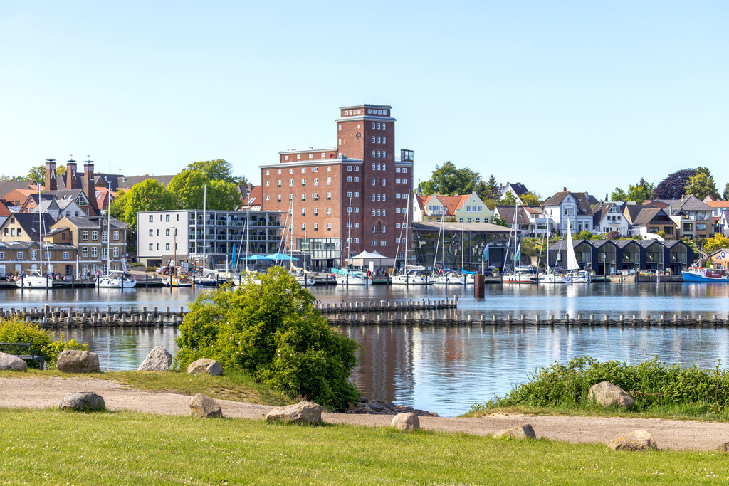 Wandbild: Hafen in Kappeln an der Schlei | Dieses Wandbild im Querformat zeigt den Hafen in Kappeln an der Schlei. Auf dem ruhigen Wasser auf der Schlei spiegeln sich die Gebäude am Hafen. Im Vordergrund befindet sich eine Rasenfläche. - Realisiert mit Pictrs.com
