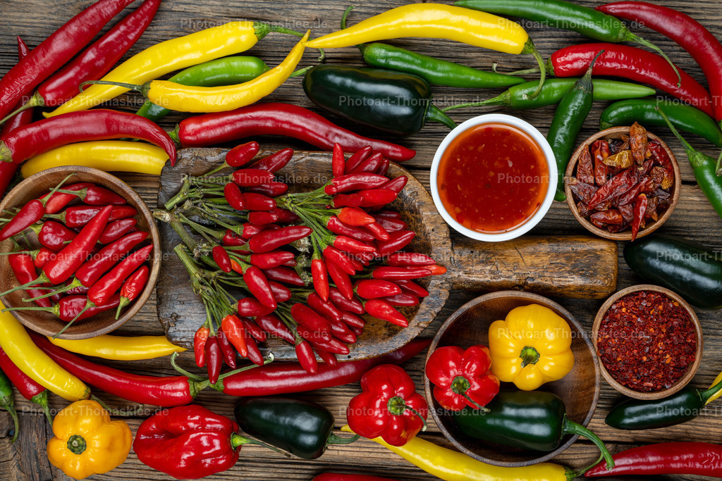 Scharfe Gewürze - Chilischoten und Paprika Vielfalt | Diese übersichtliche Anordnung zeigt eine große Vielfalt an Chili- und Paprikasorten auf verwittertem Holzuntergrund. Rote Chilischoten bilden das Herzstück der Komposition, während gelbe und grüne Paprikaschoten Farbakzente setzen. Verschiedene Holzschalen enthalten getrocknete Gewürzmischungen und Chiliflakes. Eine weiße Porzellanschale präsentiert scharfe rote Sauce. Das warme Licht betont die natürlichen Texturen und kräftigen Farben. Diese authentische Darstellung eignet sich perfekt für Kochbücher, Gewürzhändler oder kulinarische Zeitschriften und vermittelt pure Küchenkultur. - Realisiert mit Pictrs.com