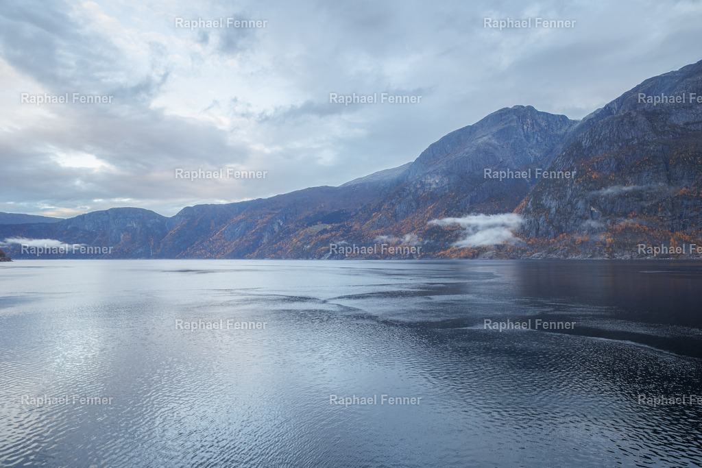 Ruhiger Morgen in Eidfjord, Norwegen | Starten Sie Ihren Tag mit der friedlichen Schönheit von Eidfjord in Norwegen, eingefangen an einem stillen Morgen während einer malerischen Kreuzfahrt. Die sanften blauen Wasserflächen verschmelzen mit den nebelverhangenen Bergen, in denen herbstliche Farben das raue Terrain zieren. Ein ideales Kunstwerk für alle, die Inspiration in der stillen Pracht der Natur finden und nordische Gelassenheit in ihren Raum bringen möchten.
