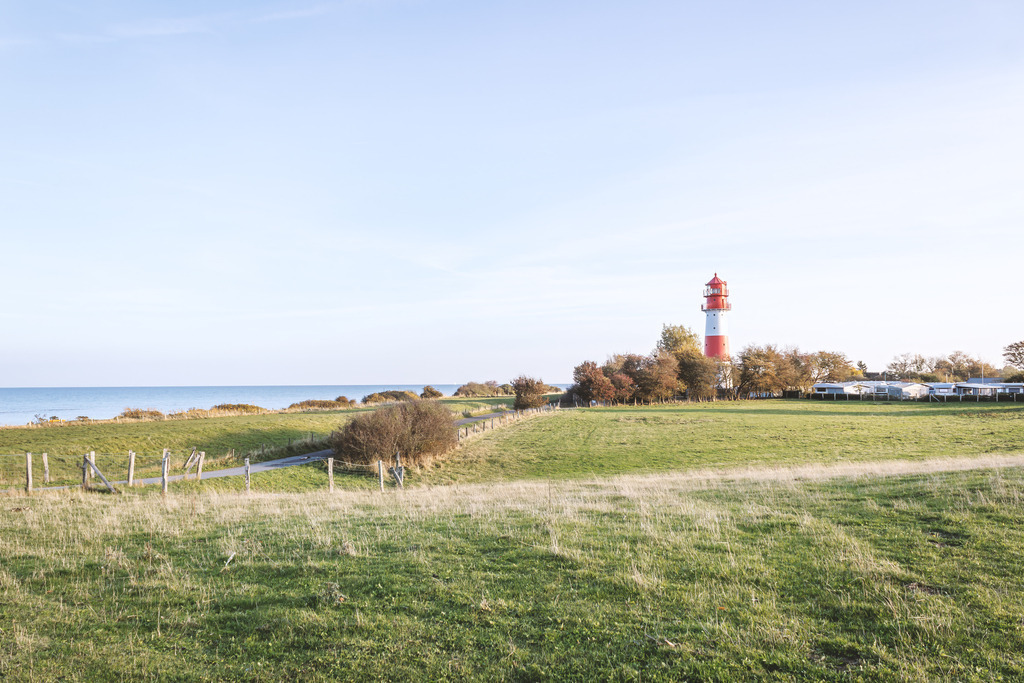 Wandbild: Weites Land Falshöft - Küstenblick und Leuchtturm | Dieses harmonische Wandbild fängt die weite und unberührte Landschaft rund um Falshöft ein. Im Vordergrund erstrecken sich grüne Weiden und Deichwiesen, die den Blick zum charakteristischen rot-weißen Leuchtturm und dem angrenzenden Küstenstreifen lenken, während links das ruhige Band der Ostsee den Horizont bildet. Die klaren Lichtverhältnisse und die weite Perspektive vermitteln ein Gefühl von Freiheit und nordischer Weite – eine stilvolle Bereicherung für anspruchsvolle Wohnräume.