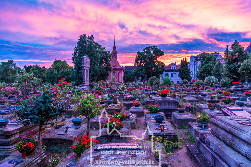 Nürnberger Stadtbild | Der Johannisfriedhof in Nürnberg bei Sonnenuntergang. Zahlreiche Gräber, die mit Blumen geschmückt sind, erstrecken sich über das Bild. Im Hintergrund erhebt sich eine kleine Kapelle, und der Himmel ist in kräftigen Orange- und Violetttönen gefärbt. Bäume und Gebäude umrahmen die Szene. - Realisiert mit Pictrs.com