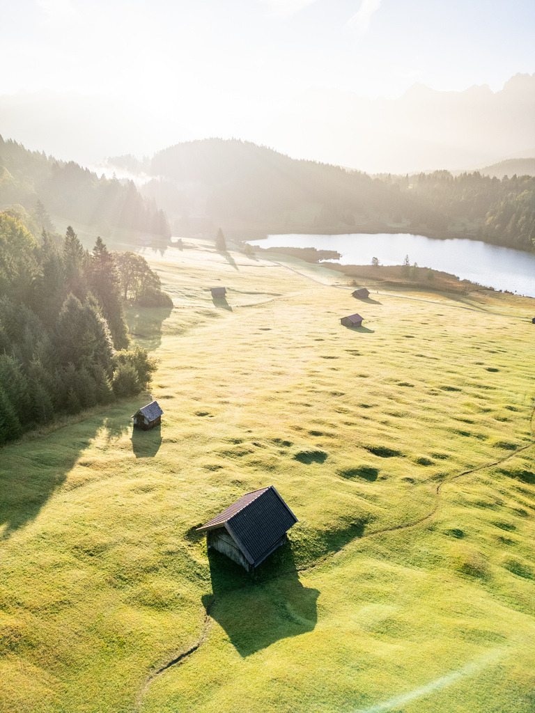 Almwiese mit Hütte im Morgenlicht | Eine kleine Hütte liegt inmitten einer weitläufigen Almwiese, während das warme Morgenlicht über die Landschaft zieht. Der See und die sanften Bergformen im Hintergrund verleihen der Szene eine ruhige, alpine Atmosphäre. - Realisiert mit Pictrs.com