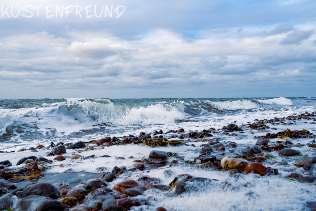 Wellen am Steinstrand - Ostsee Bilder | Dieses Wandbild zeigt kräftige Wellen, die auf den Naturstrand rollen und Seegras anspülen. Beim Zurückfließen bilden sie leichten Schaum auf den Steinen. Der Himmel präsentiert sich an diesem Tag in einem sanften Weiß-Grau. Im Wasser sind der aufgewühlte Sand vom Grund des Meeres und das Seegras deutlich zu erkennen. Ein harmonisches Küstenmotiv, das als Ostsee Bild die natürliche Schönheit der Küste eindrucksvoll einfängt.<br><br><strong>Tipp:</strong> Statt das klassische Querformat zu wählen, kannst du dieses Wandbild mit kräftigen Wellen und Naturstrand auch flexibel zuschneiden – ob als quadratisches Wandbild, Hochkantformat oder Panorama. So wird dein persönliches Ostsee Bild zum perfekten Blickfang in jedem Raum.