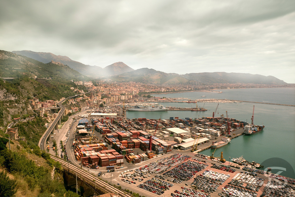 Hafen von Salerno 2006: Containerterminal und Autolager am Tyrrhenischen Meer | Eine weitläufige Luftaufnahme des geschäftigen Frachthafens von Salerno in Italien im Jahr 2006, gesehen von einer erhöhten Position. Das Bild zeigt das Hafenbecken gefüllt mit gestapelten Containern und einer großen Fläche voller Neuwagen, umgeben von der dicht bebauten Küstenstadt und bergigem Hinterland unter einem bewölkten Himmel. - Realisiert mit Pictrs.com