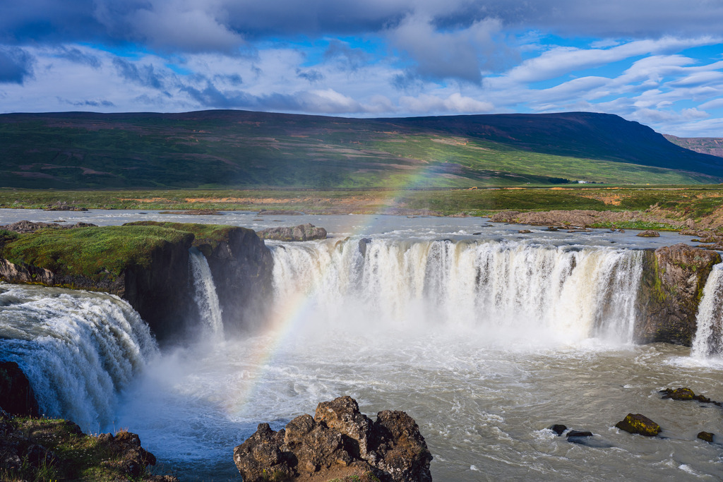 Island: Godafoss | Godafoss – der Wasserfall der Götter: Ein atemberaubendes Panorama aus tosendem Wasser, üppiger grüner Landschaft und einem strahlenden Regenbogen unter weiten Wolken. - Realisiert mit Pictrs.com