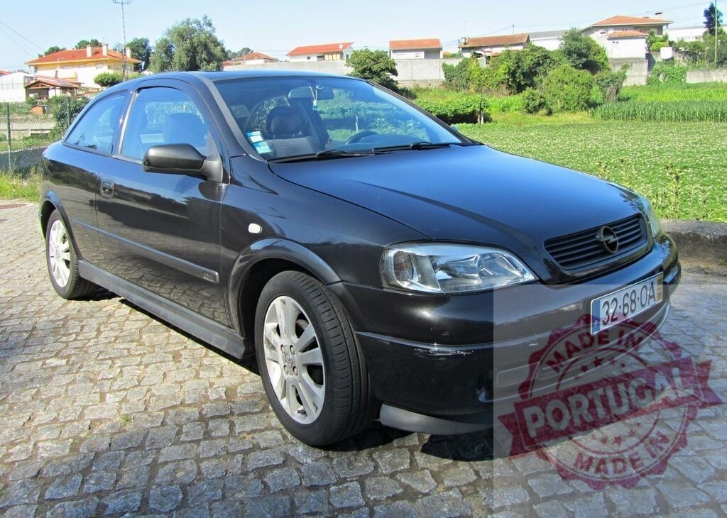 Opel Astra parked on cobblestone pavement with houses and vegetation in the background | Dark Opel Astra car parked on a cobblestone pavement, with houses and vegetation visible in the background - Realisiert mit Pictrs.com