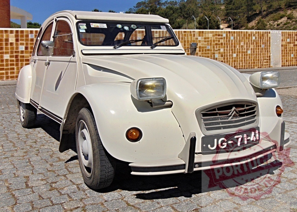 White Citroën 2CV parked on cobblestone street near tiled wall | Classic white Citroën 2CV parked on a cobblestone street, with a tiled wall in the background, on a sunny day - Realized with Pictrs.com