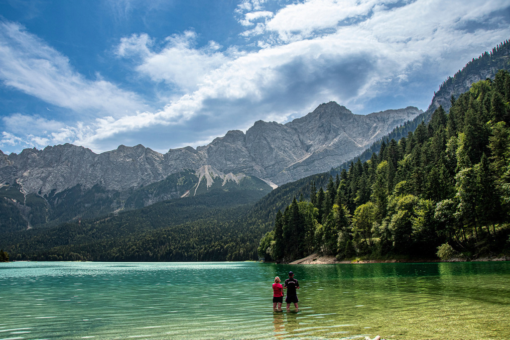 Sommermagie am Fuße der Zugspitze | Ein unvergesslicher Moment am Eibsee bei Garmisch-Partenkirchen: Ein Paar steht im glasklaren, türkisgrün schimmernden Wasser und genießt die herrliche Abkühlung an einem heißen Sommertag. Über dem dicht bewaldeten Ufer erhebt sich die imposante Felskulisse des Wettersteingebirges mit der Zugspitze unter einem dynamisch bewölkten Sommerhimmel. Die Komposition verbindet erhabene Berglandschaft mit intimer Mensch-Natur-Verbundenheit und weckt Sehnsucht nach Freiheit, Erfrischung und alpiner Idylle. - Realisiert mit Pictrs.com