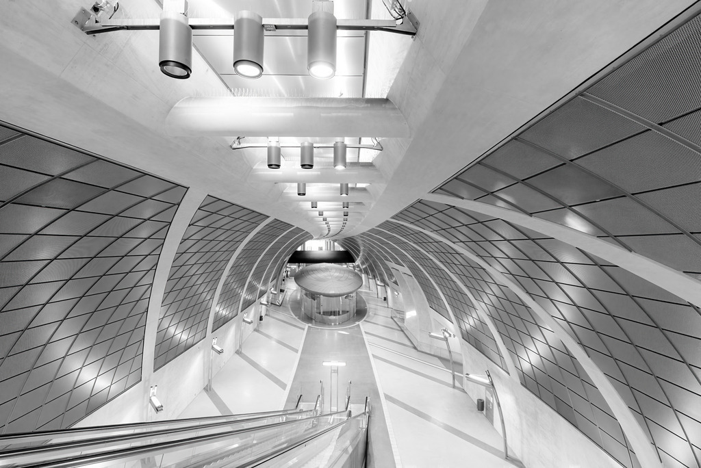 Wandbild - Futuristische Architektur einer modernen U-Bahn-Station | Das Bild zeigt die beeindruckende Architektur einer modernen U-Bahn-Station in Schwarz-Weiß. Die Perspektive, die von einer Rolltreppe aus aufgenommen wurde, betont die futuristischen, geschwungenen Linien und die symmetrischen Muster der Decken- und Wandverkleidungen. Die Beleuchtungskörper und Lüftungselemente fügen sich nahtlos in das elegante Design ein, während der Bodenbereich weit und offen wirkt. Diese minimalistische und doch hochmoderne Gestaltung vermittelt ein Gefühl von Raum und technologischem Fortschritt.
