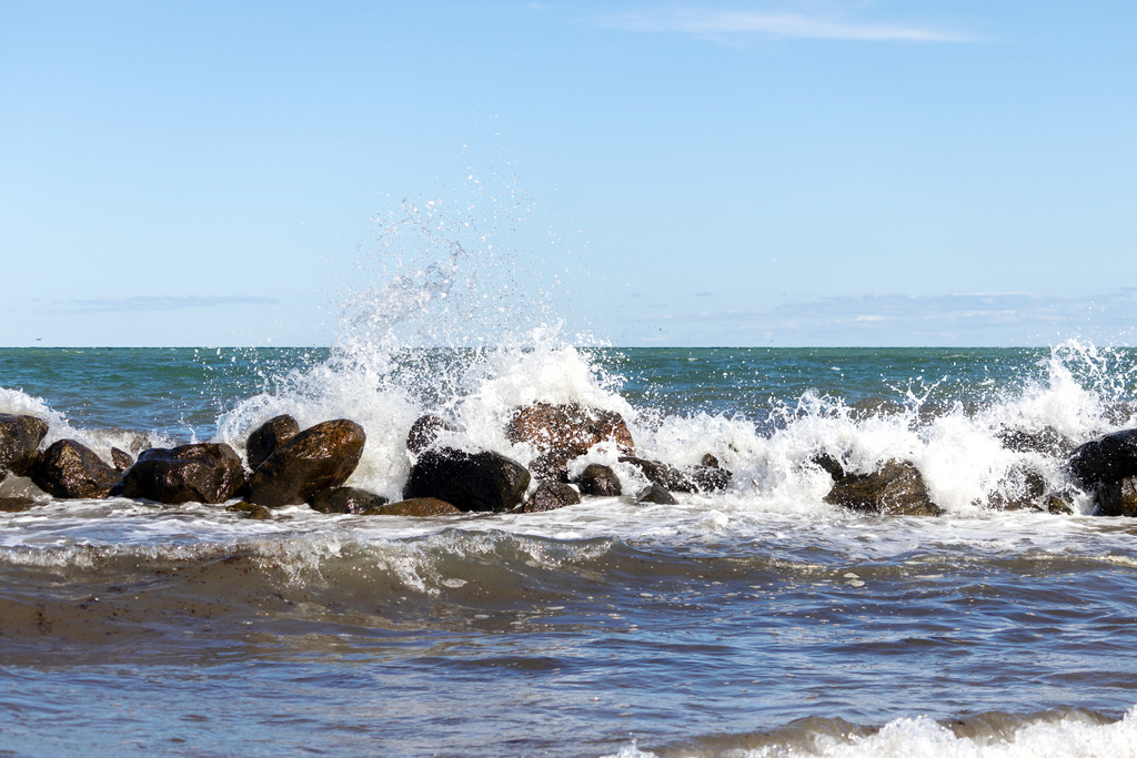 Akustikbild: Brandung am Strand in Schönhagen | Dieses Wandbild im Querformat zeigt die stürmische Ostsee im Frühling. An der Steinmole bricht eine Welle. Am blauen Himmel sind einige Wolken zu sehen.  - Realisiert mit Pictrs.com
