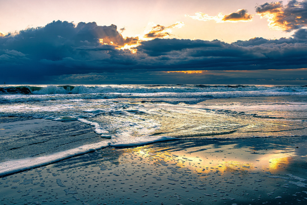 Farbenfroher Sonnenuntergang am Strand von Westerland auf Sylt | Das Bild zeigt eine Meereslandschaft bei Sonnenuntergang am Strand von Westerland auf SyltDetails der SzeneHimmel: Dunkle, schwere Wolken verdecken den Großteil des Himmels, während ein goldener Lichtschimmer im oberen Zentrum durchbricht.Meer: Eine breite Welle bricht sich im mittleren Bildbereich und erzeugt weißen Gischt.Vordergrund: Der nasse Sandstrand reflektiert das warme, goldene Licht des Himmels. - Realisiert mit Pictrs.com