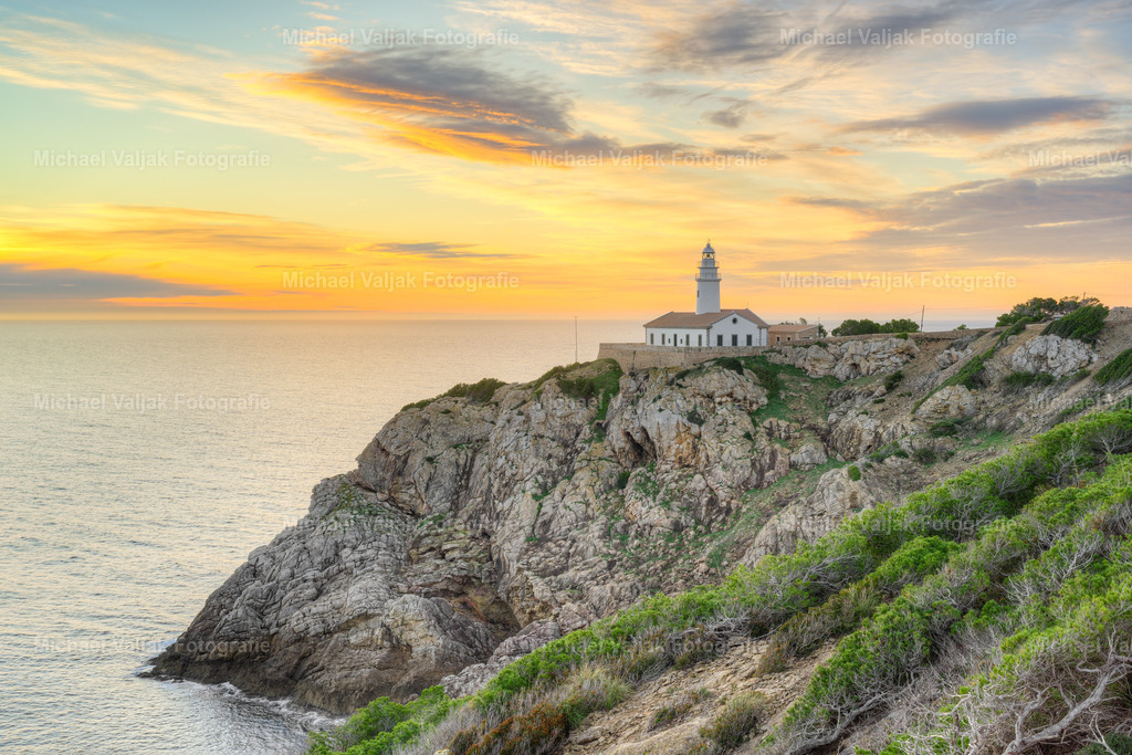 Blick zum Leuchtturm von Capdepera auf Mallorca | Der Faro de Capdepera liegt zum Sonnenaufgang in einer Landschaft, die durch ihre Felsformationen und die offene Sicht auf das Meer geprägt ist. Die warmen Farben des Himmels legen sich über die zerklüftete Küste und betonen die Formen der Klippen, die sich klar bis zur Wasserlinie abzeichnen. Der Leuchtturm steht sichtbar über der Landschaft, aber die eigentliche Wirkung entsteht durch die Küstenlinie selbst: rau, weit und vom ersten Licht weich überzogen. Ein Moment, in dem das Zusammenspiel aus Felsen, Meer und warmem Morgenhimmel die Ostküste Mallorcas besonders eindrucksvoll wirken lässt. - Realisiert mit Pictrs.com