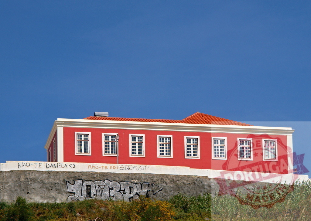 Red building with white windows on hill against blue sky | Red building with an orange roof and graffiti on a wall, under a clear blue sky - Realized with Pictrs.com