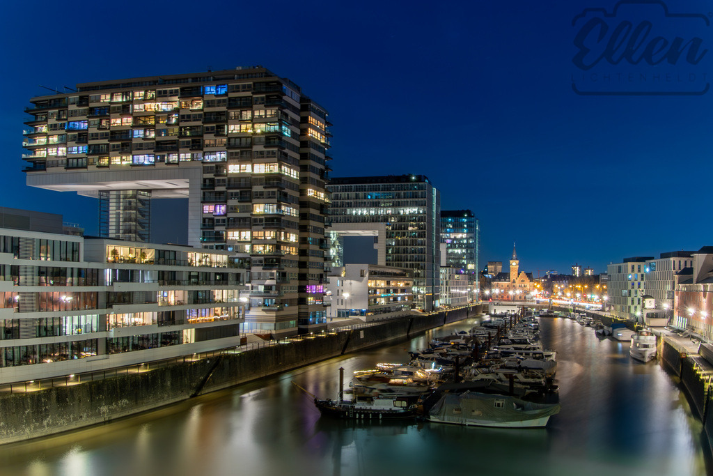 Rhythm of the Harbour | Night falls over Cologne’s Rheinauhafen, and the architecture glows like a constellation reflected in the water. The modern Kranhäuser rise above quiet boats, their mirrored lights tracing the calm rhythm of the city’s evening pulse. - Realisiert mit Pictrs.com