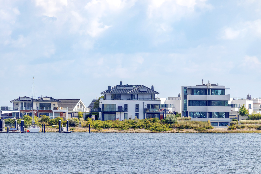 Wandbild: Ferienwohnungen in Olpenitz | Dieses Wandbild im Querformat zeigt Ferienwohnungen direkt am Meer in Olpenitz. Auf der linken Seite ist direkt vor einiger Ferienwohnung ein Segelboot zu sehen. Der Himmel ist bewölkt bei schönem Sonnenschein. - Realisiert mit Pictrs.com