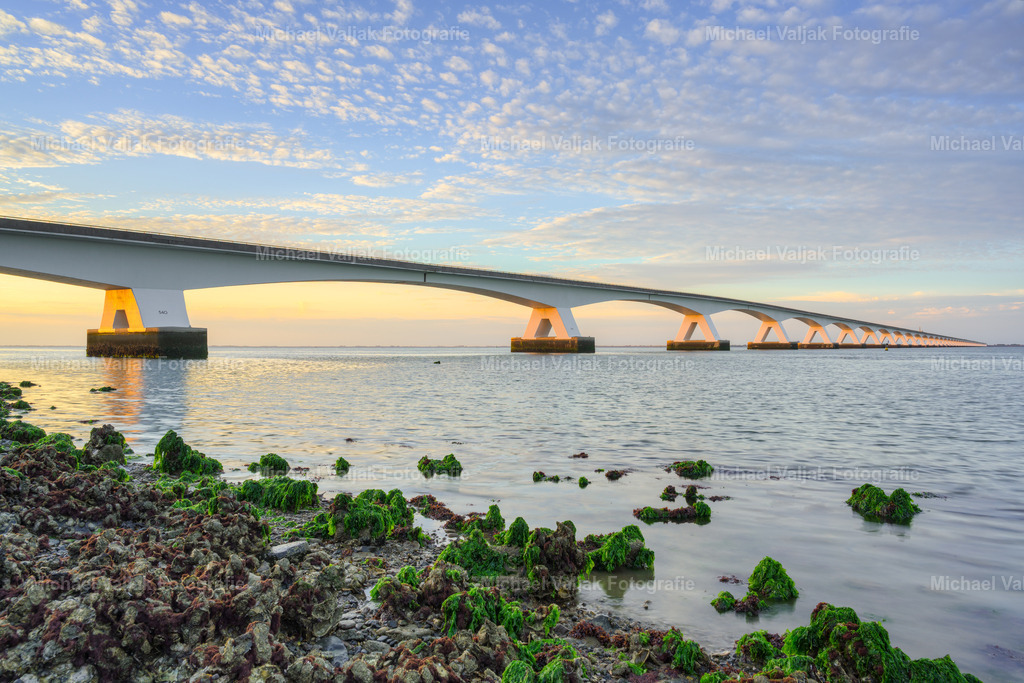 Zeelandbrücke am Abend | Vom Ufer aus fällt der Blick über die von Wasser umspülten Steine im Vordergrund hinüber zur imposanten Zeelandbrücke. Im warmen Licht des frühen Abends hebt sich die lange Reihe der Brückenpfeiler eindrucksvoll vom Himmel und der weiten Wasserlandschaft ab.Die Steine im Vordergrund verleihen dem Bild eine natürliche Tiefe und führen den Blick hinein in die Szenerie. Das sanfte Abendlicht taucht die charakteristische Brücke und die Küstenlandschaft in eine ruhige, stimmungsvolle Atmosphäre.Ein zeitloses Landschaftsfoto aus Zeeland, Niederlande, das die beeindruckende Verbindung von Natur, Wasser und Architektur an der niederländischen Küste zeigt. - Realisiert mit Pictrs.com