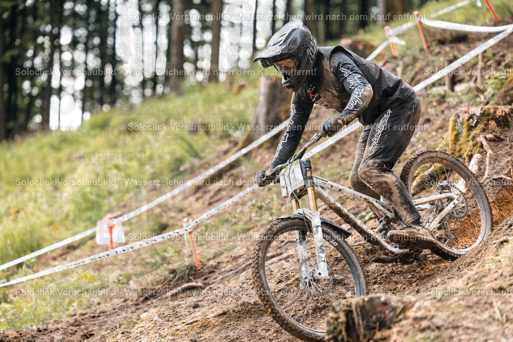 Extremsport im Gelände | Ein Mountainbiker in voller Ausrüstung rast einen steilen, schlammigen Hang hinunter. Er trägt einen schwarzen Helm, eine schwarze Brille, eine schwarze Jacke und eine schwarze Hose. Das Mountainbike ist ebenfalls mit Schlamm bedeckt und hat eine robuste Bauweise. Der Hintergrund besteht aus dichtem Wald und markierten Pfaden. - Realisiert mit Pictrs.com