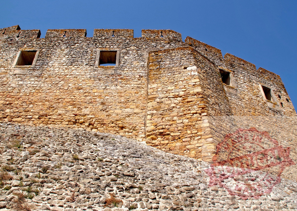 Ancient fortress stone wall architecture against clear sky | Stone fortress wall with battlements and windows against a clear blue sky - Realized with Pictrs.com
