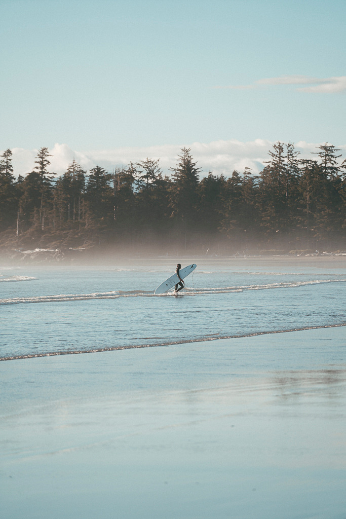 Zurück an Land – Surfermoment in Cox Bay | Ein Surfer verlässt das Wasser des Pazifiks und trägt sein Board über den feuchten Sand von Cox Bay Beach auf Vancouver Island. Die Szenerie ist ruhig, fast träumerisch – mit Nebel, der über dem Wasser liegt, und sanftem Licht, das den Moment in etwas Zeitloses verwandelt. Ein Bild voller Sehnsucht nach Freiheit und Abenteuer. - Realisiert mit Pictrs.com