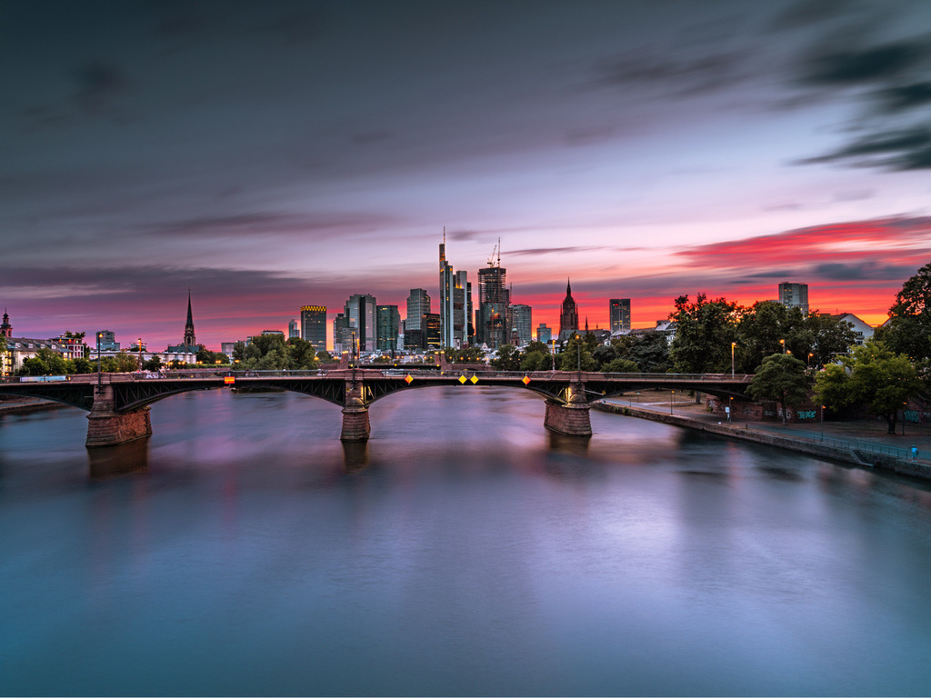 Frankfurt am Main im Abendrot | Die Frankfurter Skyline erscheint hinter einer Mainbrücke im intensiven Licht des Sonnenuntergangs. Das ruhig wirkende Wasser und der farbige Himmel verbinden Stadt und Fluss zu einem weiten Panorama. - Realisiert mit Pictrs.com
