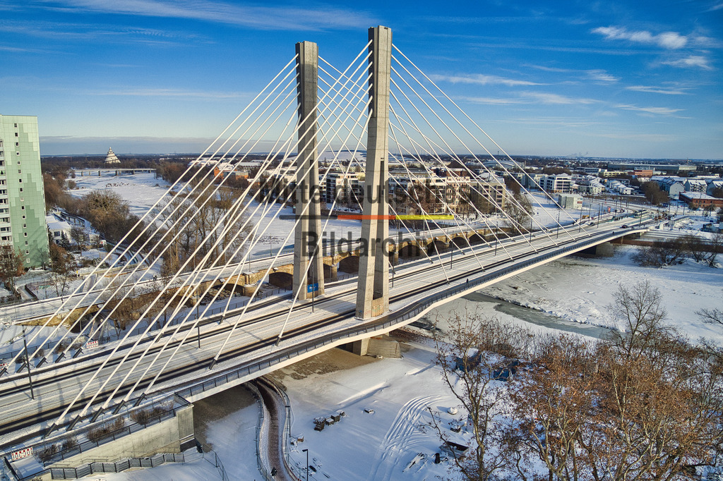 magdeburg_kaiser-otto-brücke_0007 | Das Luftbild zeigt die Kaiser-Otto-Brücke in Magdeburg aus geringer Höhe an einem winterlichen Tag. Die schneebedeckte Schrägseilbrücke hebt sich eindrucksvoll von der winterlichen Elblandschaft ab und macht die filigrane Konstruktion der Pylone und Seile besonders gut sichtbar. Aus der Drohnenperspektive werden die klaren Linien und die moderne Ingenieurarchitektur des Bauwerks eindrucksvoll hervorgehoben.Die Kaiser-Otto-Brücke wurde zwischen 2020 und 2024 als Teil des Ersatzneubaus des Strombrückenzuges errichtet. Das Bauwerk besitzt eine Gesamtlänge von 247,99 Metern und eine Breite von 27,10 Metern. Zwei 63 Meter hohe Pylone tragen den Brückenüberbau über 28 Schrägseile. Die Hauptöffnung überspannt die Alte Elbe mit einer Spannweite von 163,57 Metern. Die Brücke ist für Straßenverkehr, Straßenbahn, Radfahrer und Fußgänger ausgelegt und zählt zu den modernsten Brückenbauwerken Sachsen-Anhalts.Die winterliche Drohnenaufnahme verdeutlicht die technische Eleganz der Schrägseilkonstruktion und die präzise Ingenieurleistung des Bauwerks. Die schneebedeckte Landschaft verstärkt den Kontrast zur modernen Stahl- und Betonkonstruktion und macht die Kaiser-Otto-Brücke zu einem eindrucksvollen Beispiel zeitgemäßer Brückenbaukunst. - Realisiert mit Pictrs.com